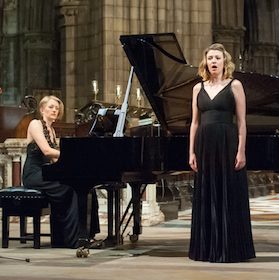 Elspeth Wyllie seated playing a grand piano, and Catherine Backhouse singing in front. Both dressed in black concert dresses, in a church.